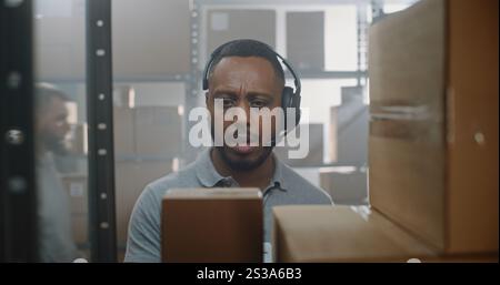 African American Inventory Specialist Talks on Headset, Checks Information Using Tablet Computer, Scans Cardboard Box, Takes Parcel for Shipping to Client. Delivery Service Sorting Center, Warehouse. Stock Photo