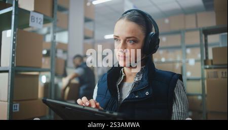Female Customer Support Specialist in Logistics in Headset Talks on Call with Client, Uses Tablet Computer. Warehouse Employees Working in Background. Delivery Service of E-Commerce Store. Close Up. Stock Photo