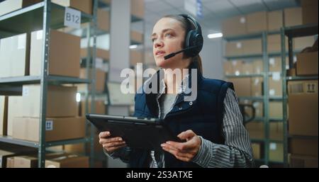 Female Logistics Coordinator, Manager Wearing Headset Chats to Customer, Uses Tablet Computer in Warehouse Facility. Sorting Center Worker Preparing Parcels with E-Commerce Online Orders. Dolly Shot. Stock Photo