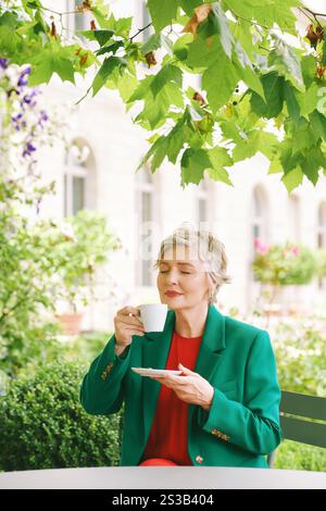 Outdoor portraot of beautiful 55 - 60 year old woman enjoying morning beverage in spring garden, wearing red green outfit, holding cup of tea or coffe Stock Photo