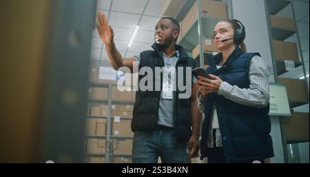 Diverse Sorting Center Employees in Warehouse Facility of E-Commerce Store: Female Manager in Headset Uses Smartphone, Scans Cardboard Boxes with Online Orders, Checks Inventory, Talks with Colleague. Stock Photo