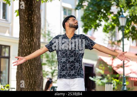 Indian young man taking a deep breath of fresh air, relaxing, taking a break, resting, meditating, calm down outdoors. Hispanic guy enjoying happy day stands on urban city street park. Town lifestyles Stock Photo