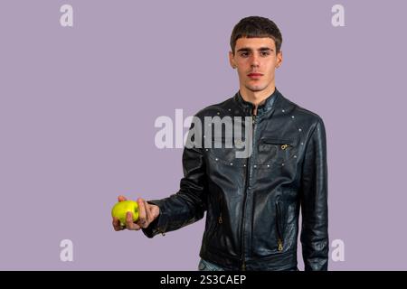 Young man wearing black leather jacket offering a green apple on neutral background Stock Photo