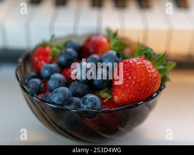 Close-up of fresh strawberries and blueberries in a black bowl with a blurred piano keyboard in the background. Vibrant colors emphasize the freshness Stock Photo