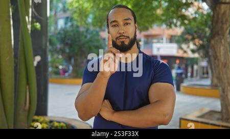 Young man with beard wearing blue shirt stands in urban park with trees and buildings blurred in the background pointing upward with a thoughtful expr Stock Photo