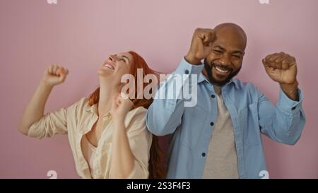 Interracial couple celebrates joyfully together against a pink background with a man in blue shirt and a woman in beige clothing. Stock Photo