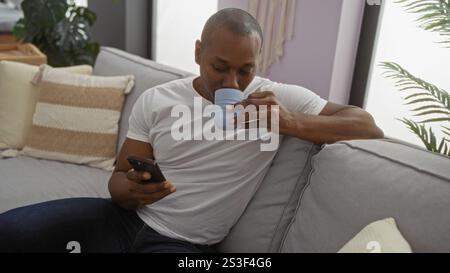 Young man relaxing on sofa sipping coffee and using smartphone in cozy living room setting with modern decor and sunlight filtering through large wind Stock Photo