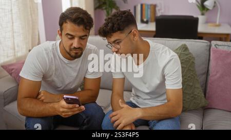 Hispanic father and son bonding over a mobile phone on a cozy couch in a living room, showcasing family love and togetherness in an indoor setting. Stock Photo