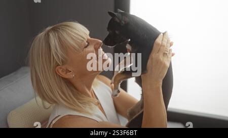 Mature blonde woman smiles while holding a small chihuahua dog in the living room of her apartment, exhibiting a heartwarming indoor moment. Stock Photo