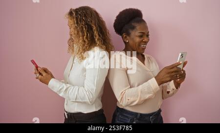 Women friends using phones together, standing against an isolated pink wall background, depicting two adult females connecting and interacting. Stock Photo