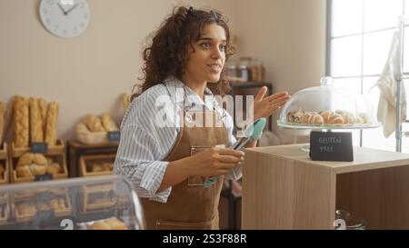 Young woman working in a bakery setting indoors, demonstrating gluten-free products while smiling and wearing an apron. Stock Photo