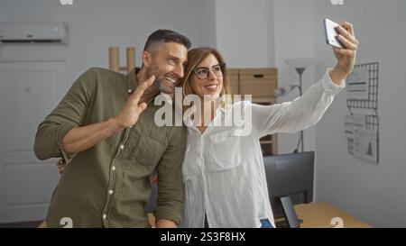 Employees taking a selfie during a video call in an office, smiling man and woman standing close together indoors Stock Photo