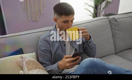 Young man in a home setting sips coffee while browsing a smartphone on a cozy couch in the living room. Stock Photo