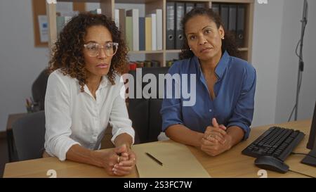 Latin women in an office setting, discussing business matters while sitting at a desk with office supplies in the background. Stock Photo