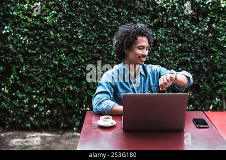 Smiling young man with curly hair sitting outdoors at a red table, working on a laptop, checking his watch, with a smartphone and coffee cup nearby, g Stock Photo