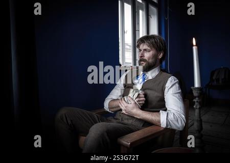 A solemn man in traditional vintage clothing sits in a blue-walled room, holding an old book, with a burning candle casting a soft light, creating a n Stock Photo