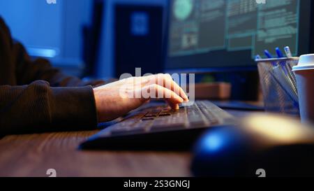 Hacker in secret underground shelter typing on keyboard, writing lines of code on computer, trying to get past security systems with virus. Scammer working on hacking script, camera A close up shot Stock Photo