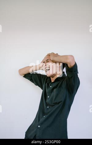 A man in a black shirt expressing frustration or stress, holding his head with both hands and shouting, standing against a plain white background Stock Photo