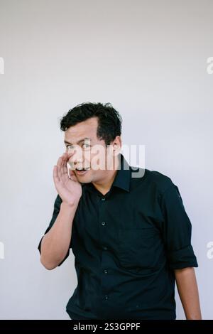 A man dressed in a black shirt making a playful gesture, whispering with his hand near his mouth against a plain white background. Perfect for concept Stock Photo