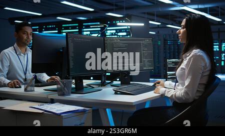 Team of technicians in data center designing, implementing and managing infrastructure to ensure optimal performance and reliability. Teamworking colleagues in server room coding on computer, camera B Stock Photo