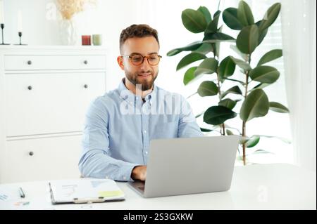 Attractive successful arabian or caucasian man with glasses in a blue shirt, company manager or IT specialist, working online using a laptop on a project while sitting at a work desk, smiling Stock Photo