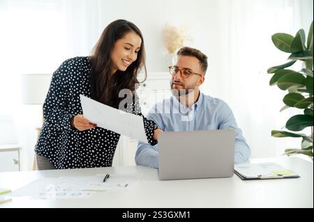 Professional woman holding a report and presenting it to a man with glasses, they discussion ideas while sitting at a desk with a laptop and data sheets in a contemporary workspace, smiling Stock Photo