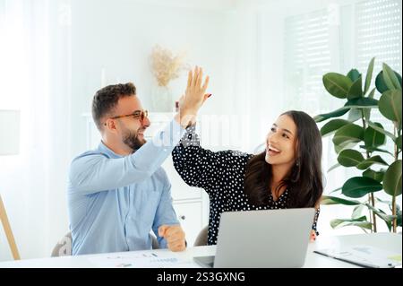 Cheerful multiracial colleagues giving each other a high five in celebration of a successful project, surrounded by graphs and a laptop, capturing a moment of joy and accomplishment in the workplace Stock Photo