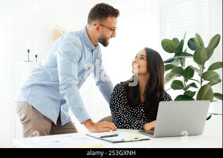 Smiling arabian or indian woman in sits at a desk with a laptop, looking up at a confident man in glasses in the office, multiracial business colleagues working together on a project, discussing ideas Stock Photo