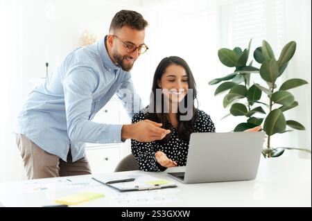 Professional young man in glasses leaning over a smiling brunette woman, pointing at a laptop screen during a business discussion in a modern office, teamwork concept, positive colleagues Stock Photo