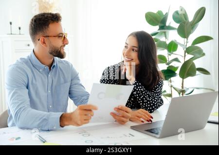 Cheerful business partners sitting side by side at a desk, with a laptop in front, analyzing data and reports on a printed sheets, discuss ways to improve results, new ideas and strategies, smile Stock Photo