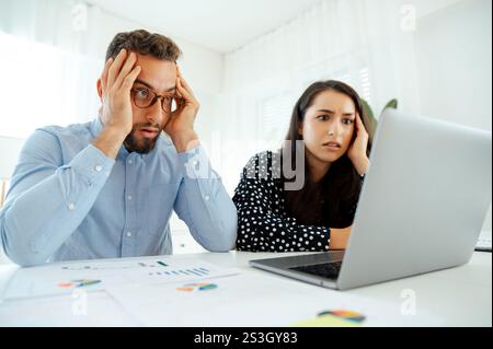 Shocked young company colleagues at a modern office, analyzing unexpected data on a laptop, with charts and graphs on the table, expressing concern and disbelief, are perplexed Stock Photo