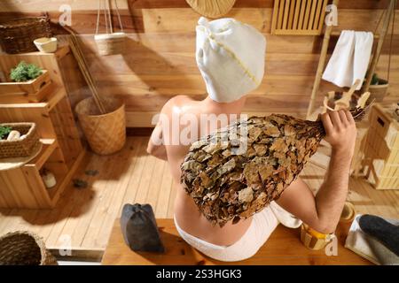 Young man with whisk sitting on bench in sauna, back view Stock Photo