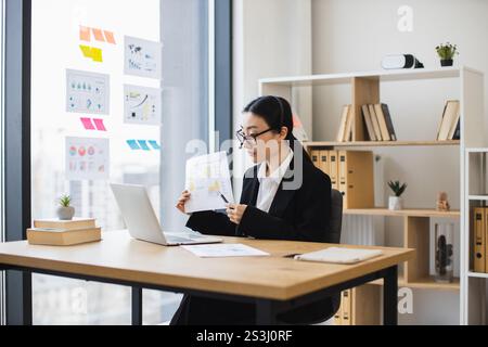 Confident Asian businesswoman wearing headset presenting data during video call using laptop. Middle-aged woman working remotely in office setting showcasing professional skills and communication. Stock Photo