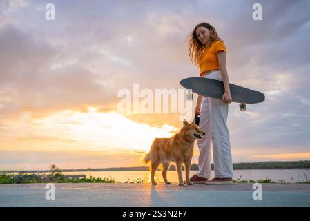 Young woman with long brown hair, wearing a yellow crop top and white pants, stands with her karelian bear dog and her longboard at sunset by a river, enjoying the beautiful golden hour light Stock Photo