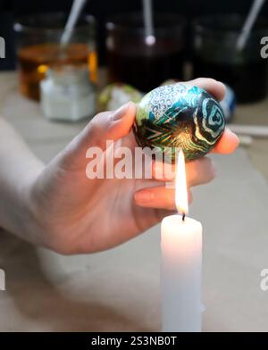 Photo of making Easter eggs. Process of removing wax from painted egg. Woman's hand holds an egg over candle flame. Wax is partially removed from the Stock Photo