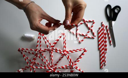 Hand made. Female hand holds needle with thread. In the process of making Christmas stars from paper tubes for cocktails. Tubes in red and white strip Stock Photo