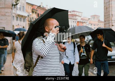 A man takes pictures and explore the city on a rainy day on a crowded street. The photographer is with the umbrella. Verona, Italy - 06 July 2024 Stock Photo