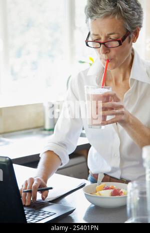 Woman Drinking Smoothie whilst Typing on Laptop Stock Photo