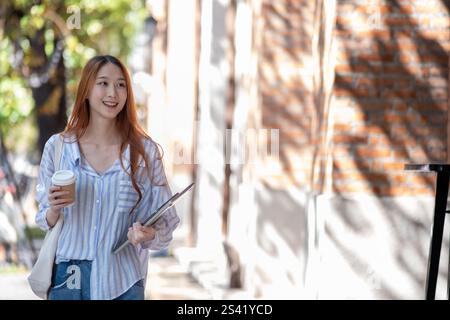 Woman is walking down the street holding a cup of coffee and a book. She is smiling Stock Photo