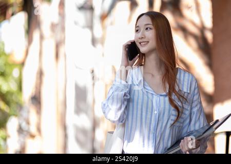 A smiling young woman talking on a mobile phone while holding a tablet and tote bag in an outdoor setting. Sunlight streams through trees, creating a Stock Photo