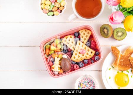 Cute Easter style lunchbox. Creative kids school lunch box with Easter symbols and sweets - bunny rabbit belgian waffle sandwiches, berry fruit, candy Stock Photo