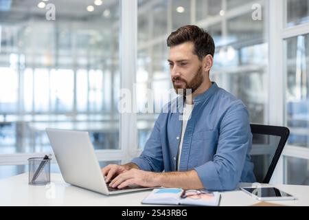 Serious and focused man typing on laptop keyboard. Businessman in casual clothes working inside office at workplace. Stock Photo