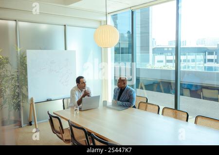 Two business executives engage in a focused discussion in a modern office setting. The room features a large window overlooking the city, natural ligh Stock Photo