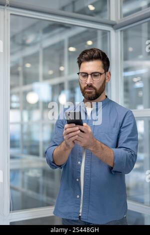 Serious confident man with phone in hands, businessman standing near window inside office at workplace, using app on smartphone, typing text message. Stock Photo