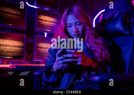 Asian woman in front of a computer monitor display using mobile phone, neon light, Hacker wear black Stock Photo
