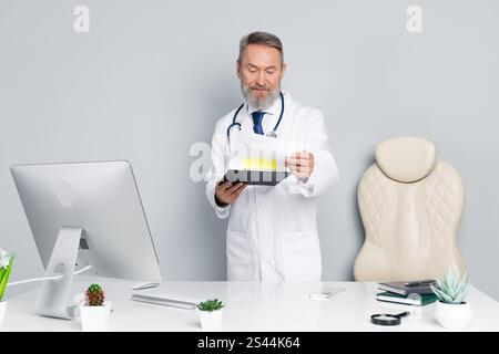 Senior male doctor in white coat reviewing medical documents at workplace in bright office Stock Photo