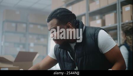 African American Warehouse Clerk Packs Cardboard Box with Online Order, Scans Code, Uses Tablet Computer, Prepares Parcels for Shipping. Sorting Center of E-Commerce Store, Delivery Service. Close Up Stock Photo