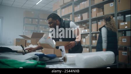 African American Warehouse Clerk Puts Sticker on Cardboard Box with Online Order, Prepares Parcels for Shipping, Scans Code, Uses Tablet Computer. Sorting Center of E-Commerce Store, Delivery Service. Stock Photo