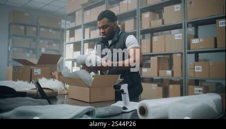 African American Warehouse Clerk Puts Sticker on Cardboard Box with Online Order, Prepares Parcels for Shipping, Scans Code, Uses Tablet Computer. Sorting Center of E-Commerce Store, Delivery Service. Stock Photo