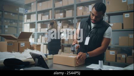 African American Warehouse Clerk Puts Sticker on Cardboard Box with Online Order, Prepares Parcels for Shipping, Scans Code, Uses Tablet Computer. Sorting Center of E-Commerce Store, Delivery Service. Stock Photo